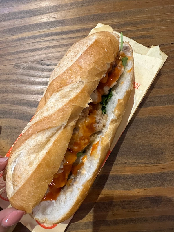 A person's hand holding a crispy Vietnamese Bánh Mì sandwich. The baguette is filled with savory meat, chili sauce, and cilantro, resting on a wooden table.
