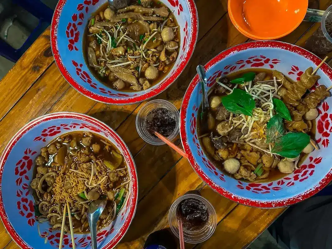 Three bowls of authentic Thai Boat Noodles (Kuay Teow Reua) with rich, dark broth and meatballs served in traditional enamel bowls.