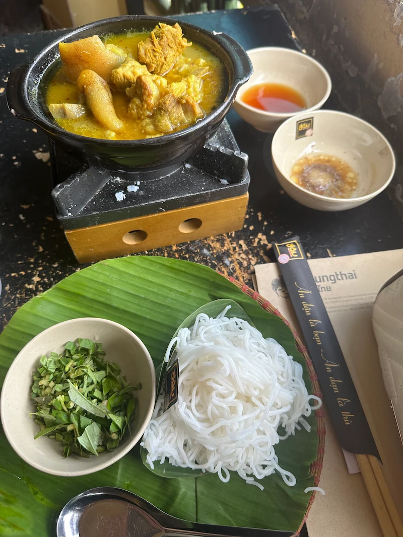 A Vietnamese curry meal served deconstructed. A black pot of yellow pork curry sits on a small stove, next to a plate with fresh rice vermicelli noodles and herbs.