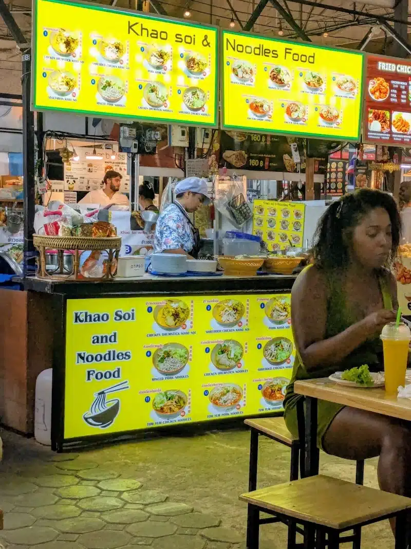 A bright yellow menu board at a street food stall advertising "Khao Soi & Noodles," with a tourist enjoying a meal in the foreground.