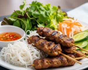 A plate of Vietnamese grilled pork skewers (Nem Lụi or Nem Nướng). The charcoal-grilled meat is served with a side of rice vermicelli noodles, a large pile of fresh herbs, and a small bowl of nước chấm dipping sauce.