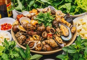 A large platter overflowing with a variety of cooked snails (ốc), clams, and mussels. The communal seafood dish is surrounded by fresh Vietnamese herbs, basil, and bowls of chili.
