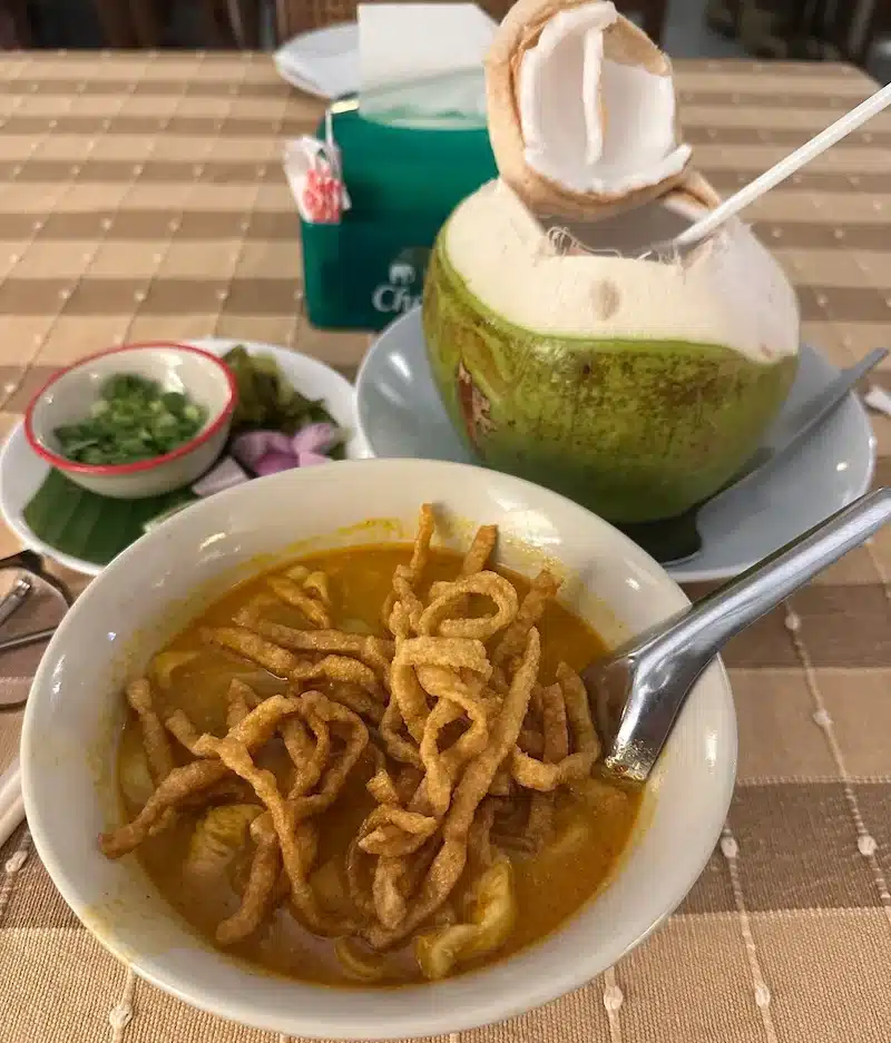 A bowl of rich Northern Thai Curry Noodles (Khao Soi) topped with crispy noodles, served alongside a fresh young coconut drink.