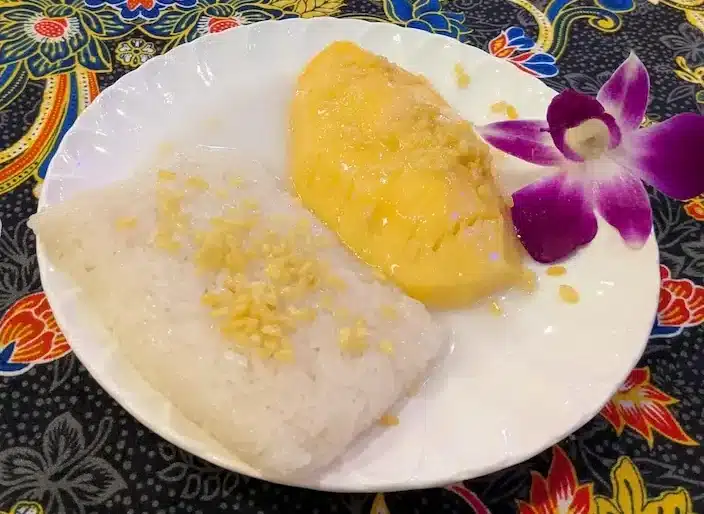 A serving of Mango Sticky Rice (Khao Niao Mamuang), the famous Thai dessert, topped with crispy mung beans and garnished with an orchid.