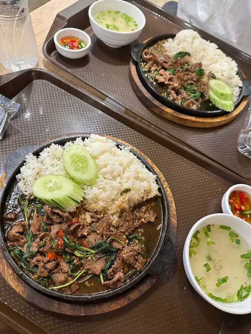 Two sizzling hot plates of Pad Kra Pao (Holy Basil Stir-Fry) served with jasmine rice and cucumbers, a popular spicy lunch option in Thailand.