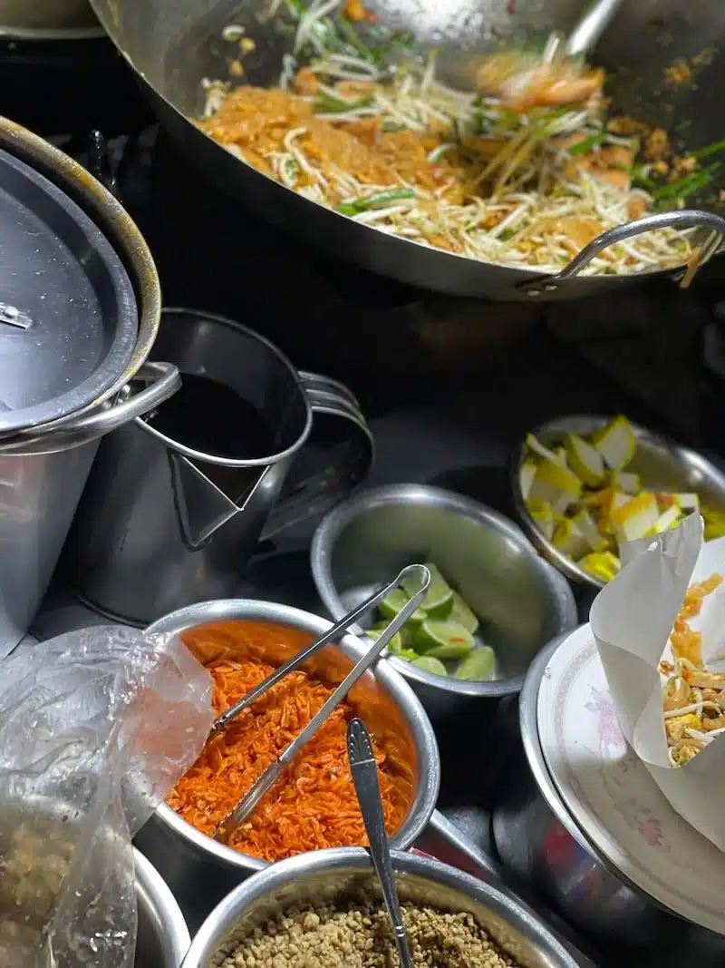 A street food vendor cooking fresh Pad Thai in a large wok, surrounded by ingredients like dried shrimp, tofu, and lime wedges.