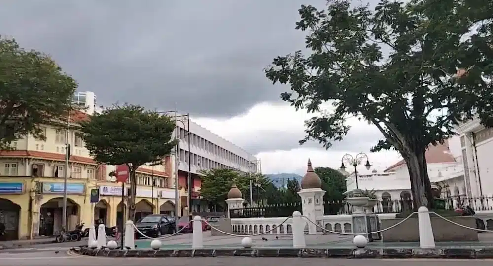A street corner in Georgetown, Penang, showing a mix of colonial-era shophouses and modern buildings under a cloudy sky. The architecture is typical of the UNESCO World Heritage site.