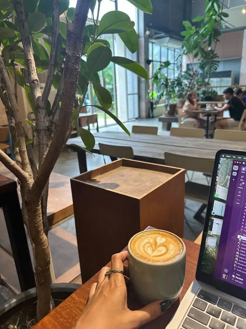 A digital nomad working on a laptop while holding a hot latte at a trendy, plant-filled cafe in Chiang Mai, highlighting the city's popular remote work culture.