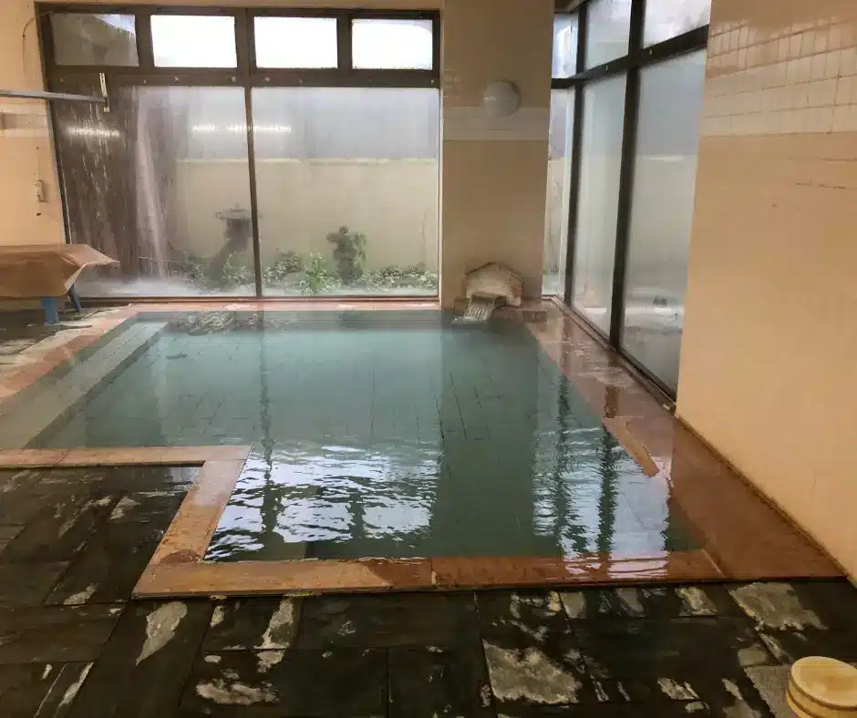 An empty indoor Japanese onsen bath with a large tiled pool and garden view, highlighting the clean and peaceful atmosphere where strict bathing etiquette applies.