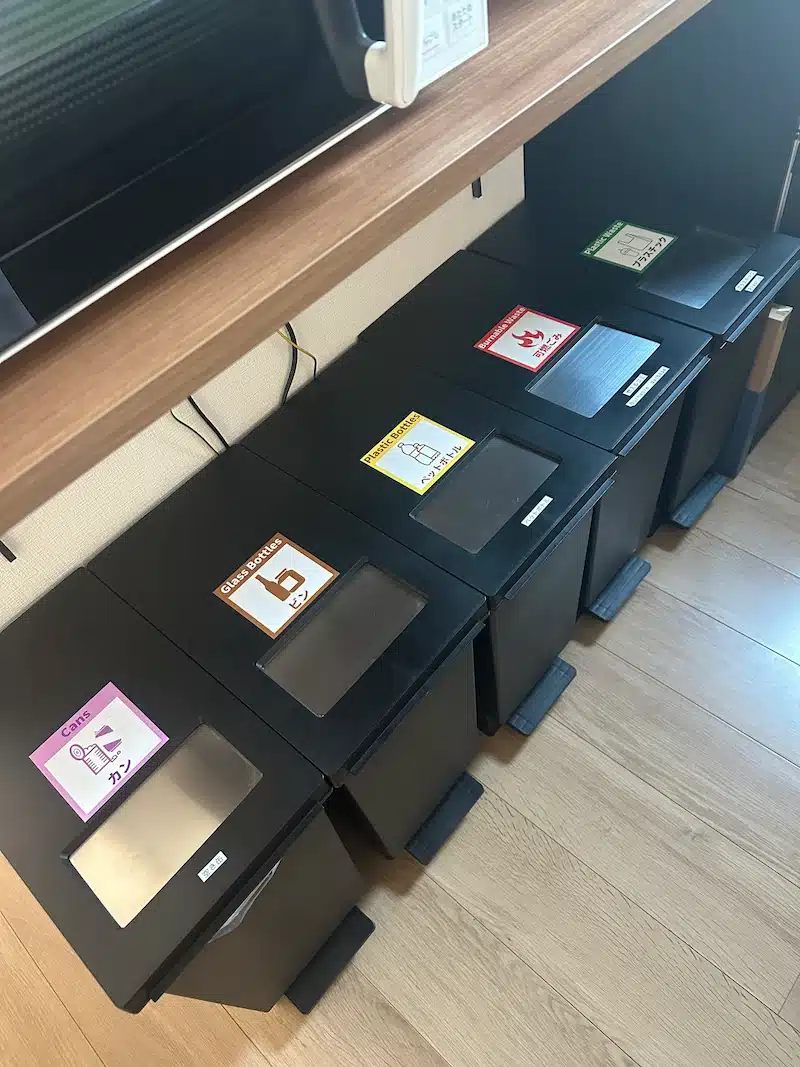 A row of color-coded recycling bins in Japan clearly labeled for cans, bottles, and burnables, demonstrating the strict waste separation rules tourists must follow.