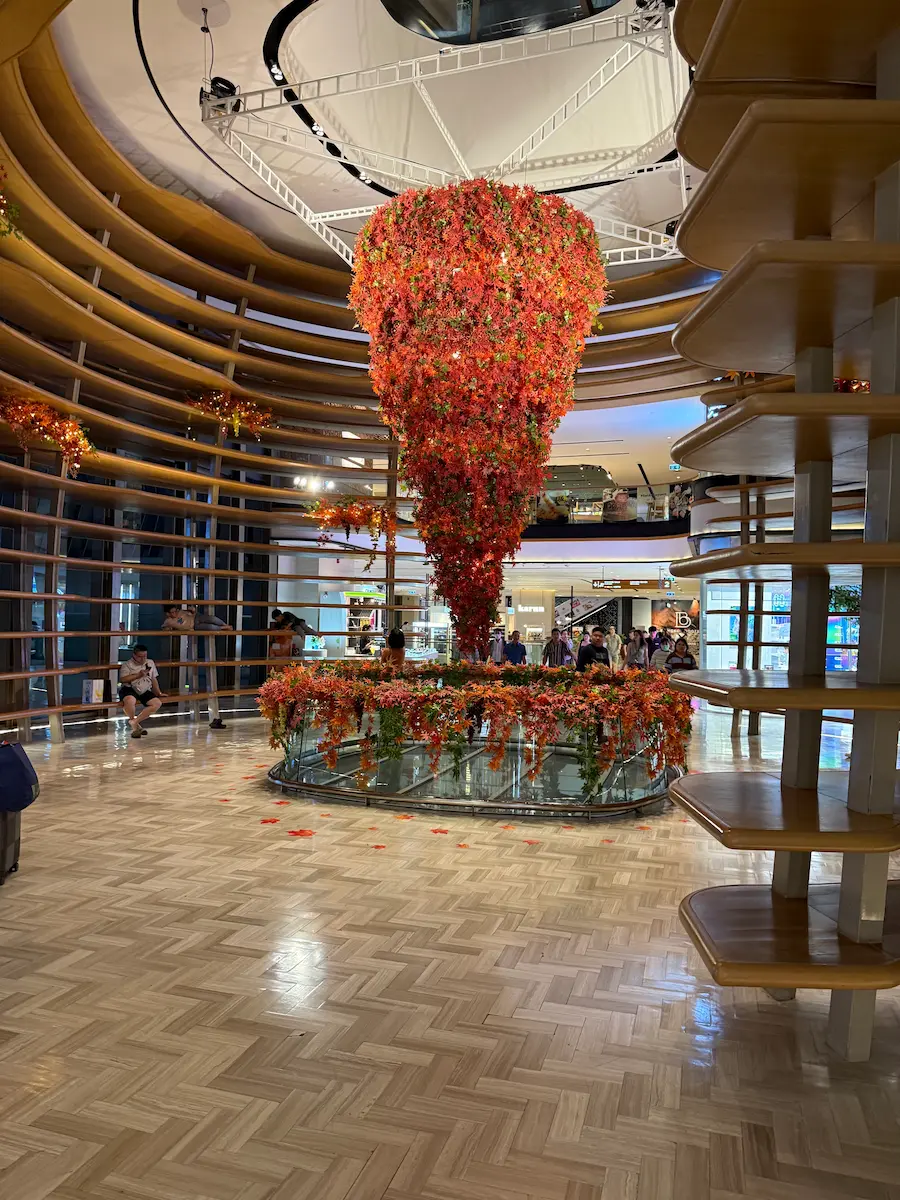 A stunning, cone-shaped art installation made of thousands of red and orange flowers hangs in the center of a modern, circular atrium in a Bangkok mall.