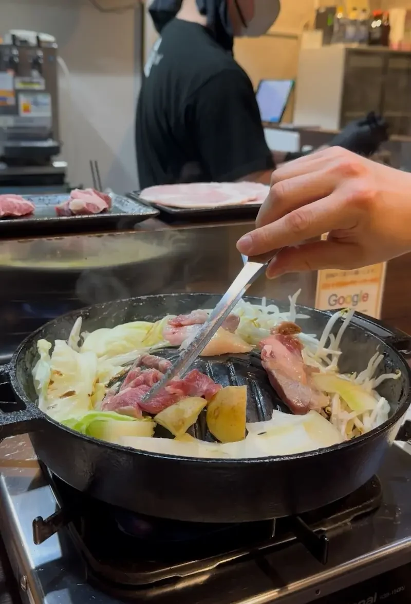 A diner is grilling Jingisukan (Japanese grilled lamb) on a convex cast-iron skillet set on a tabletop gas stove. Tongs are used to move the meat amongst sizzling cabbage, bean sprouts, and onions.