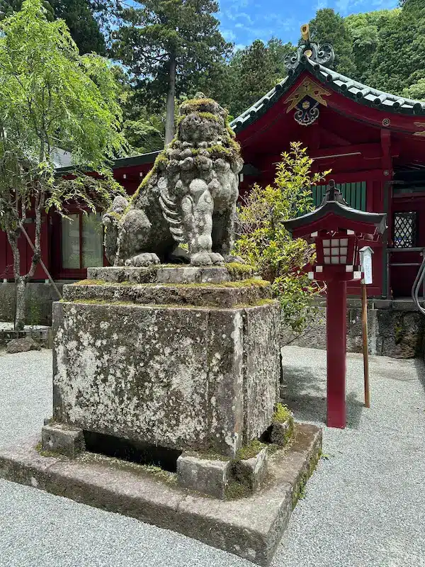 A moss-covered Komainu guardian lion-dog statue sitting in front of a traditional red Shinto shrine in Japan, symbolizing the protection of sacred grounds.