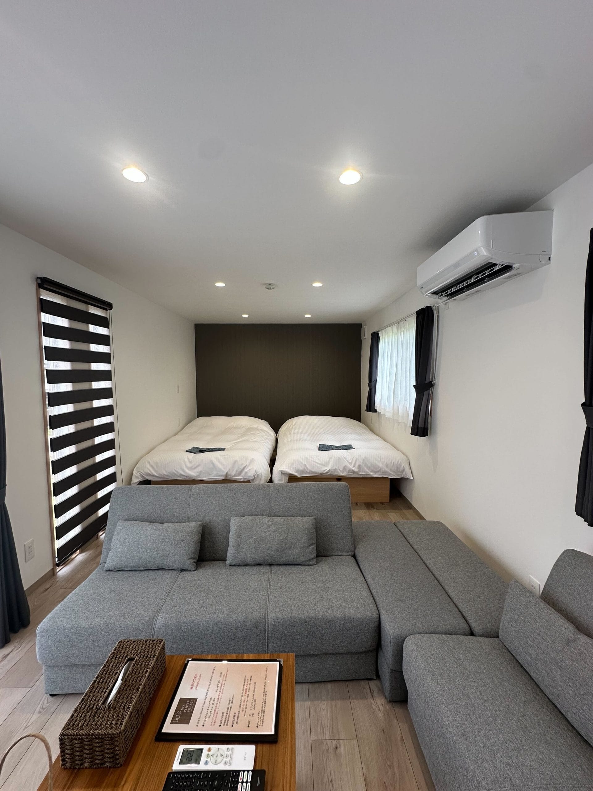 The clean and modern interior of the cabin-style trailer accommodation at Lake Toya, showing two beds pushed together and a comfortable grey sectional sofa.