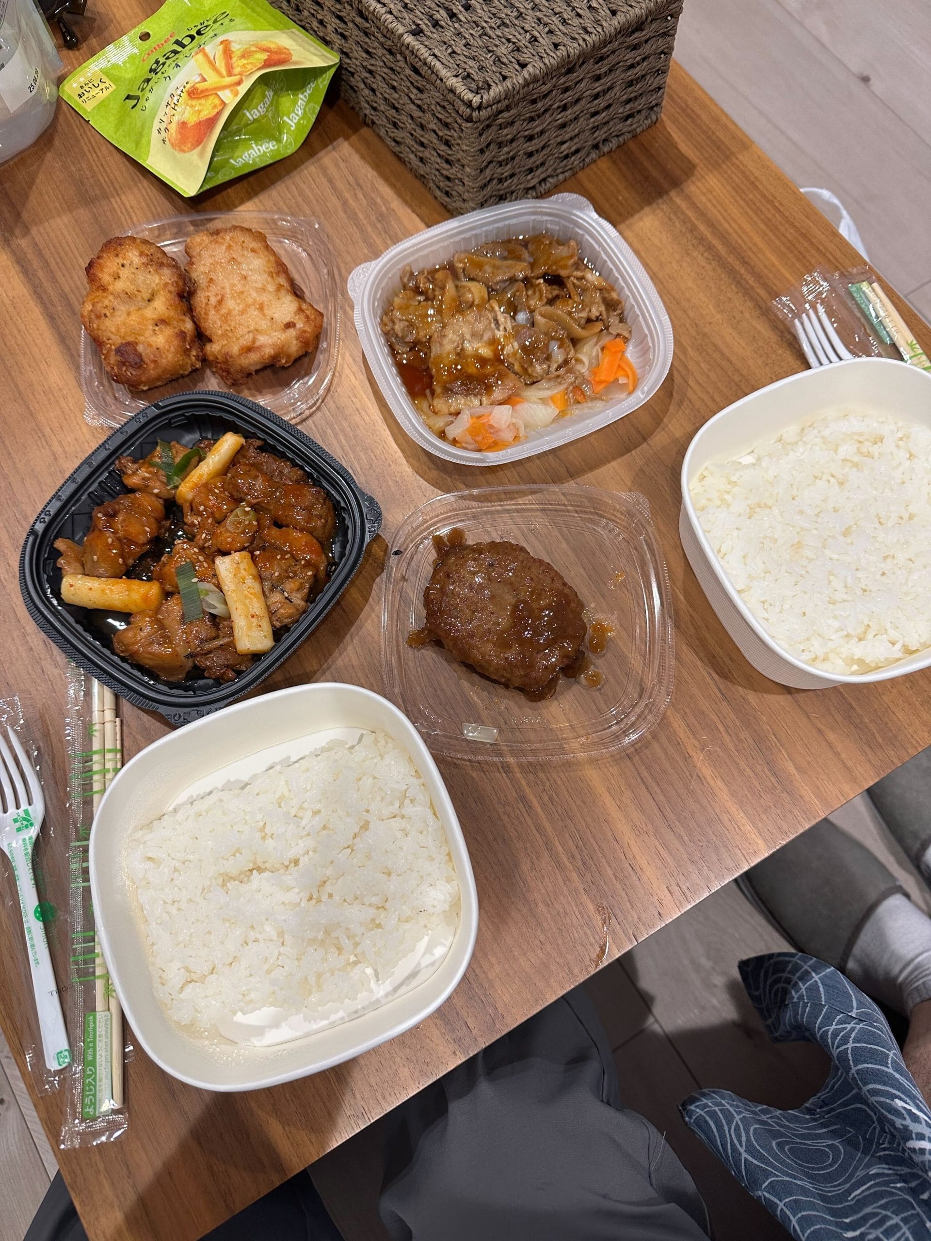 An affordable and delicious dinner from a Japanese convenience store, featuring various dishes like sweet and sour pork, a hamburger patty, beef bowl, and white rice laid out on a wooden table.