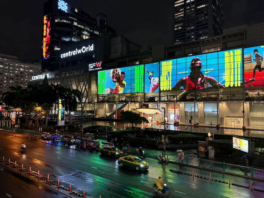An impressive nighttime view of the CentralWorld shopping mall in Bangkok. The entire facade is illuminated by massive, vibrant LED screens, with the wet street below reflecting the colorful lights.