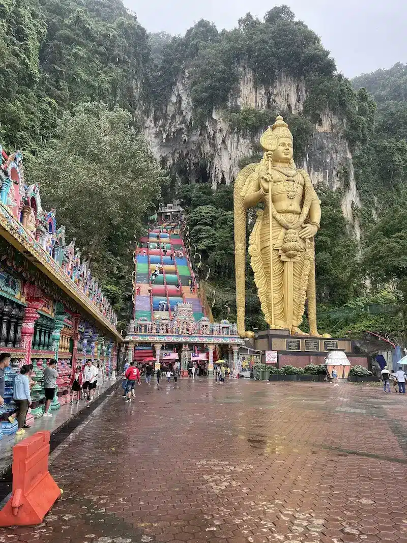 The massive golden statue of Lord Murugan standing next to the vibrant, rainbow-colored staircase at Batu Caves. The wet ground reflects the colorful entrance of this iconic Hindu temple.