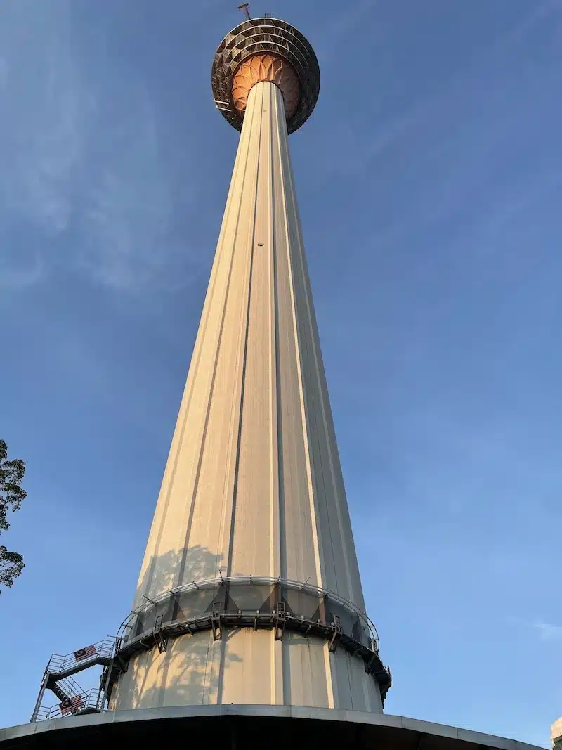 Looking straight up at the concrete shaft and bulbous observation deck of the KL Tower (Menara KL). The telecommunications tower stands tall against a clear blue sky.