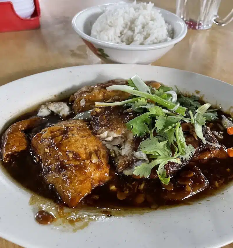 A plate of deep-fried fish fillets smothered in a savory dark soy sauce and topped with fresh coriander. Served with a bowl of steamed white rice at a local Malaysian restaurant.