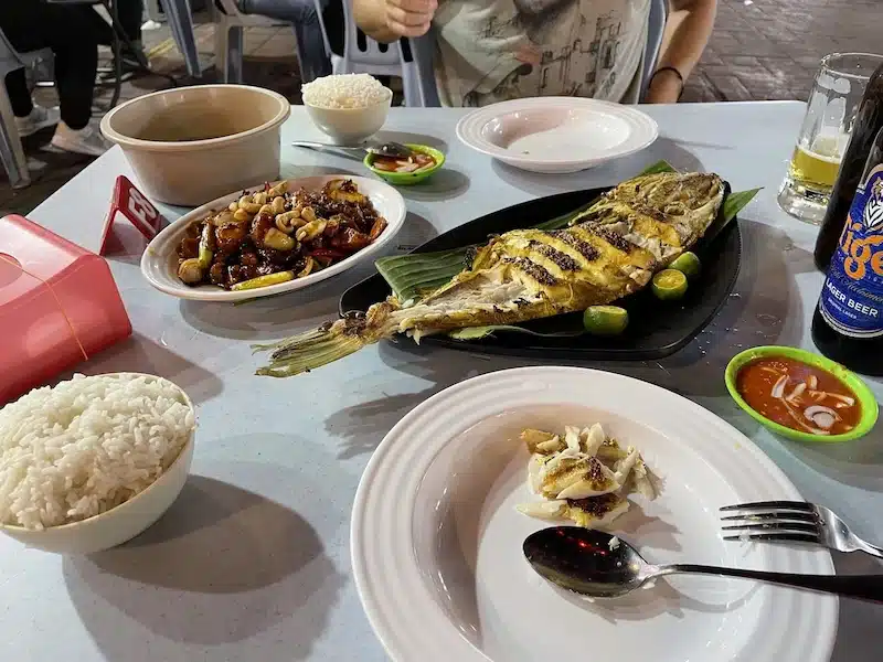 A classic Malaysian street food feast featuring grilled fish (Ikan Bakar) served on a banana leaf. The table is set with stir-fried seafood, rice, and a bottle of Tiger beer.