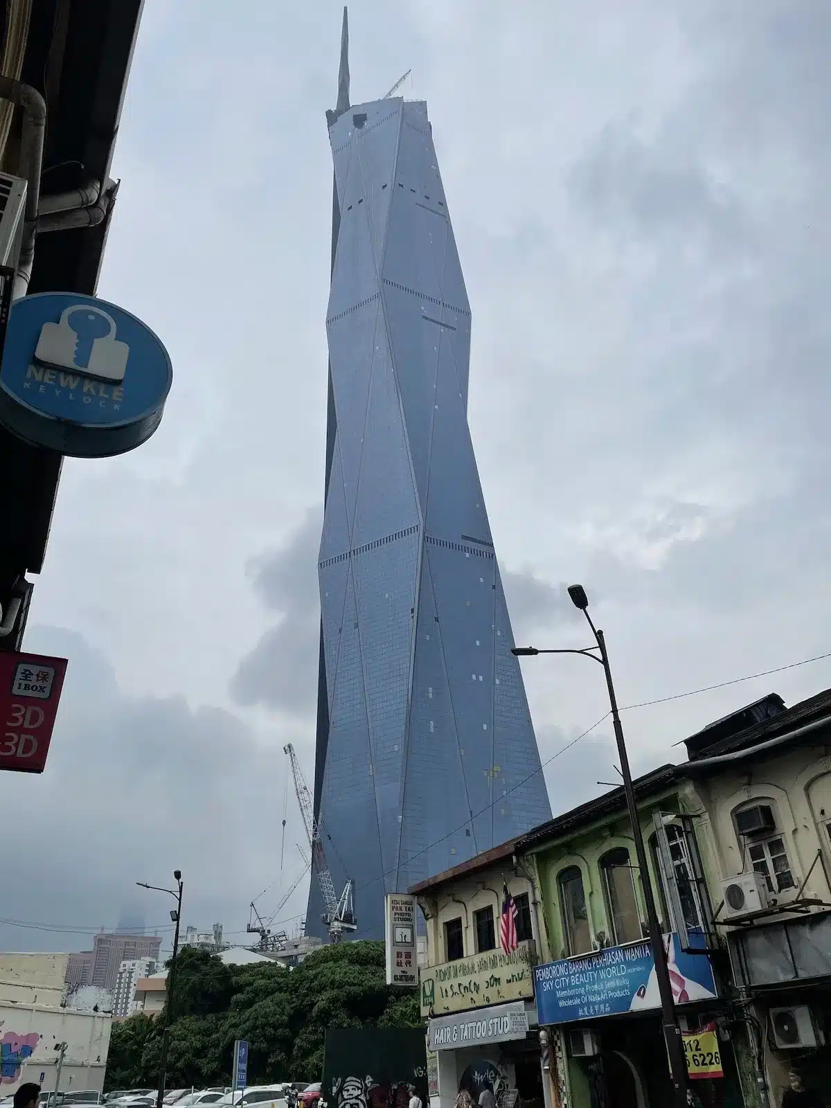 The futuristic Merdeka 118 skyscraper towering over traditional heritage shophouses near Chinatown. The modern glass facade contrasts sharply with the historic architecture below.