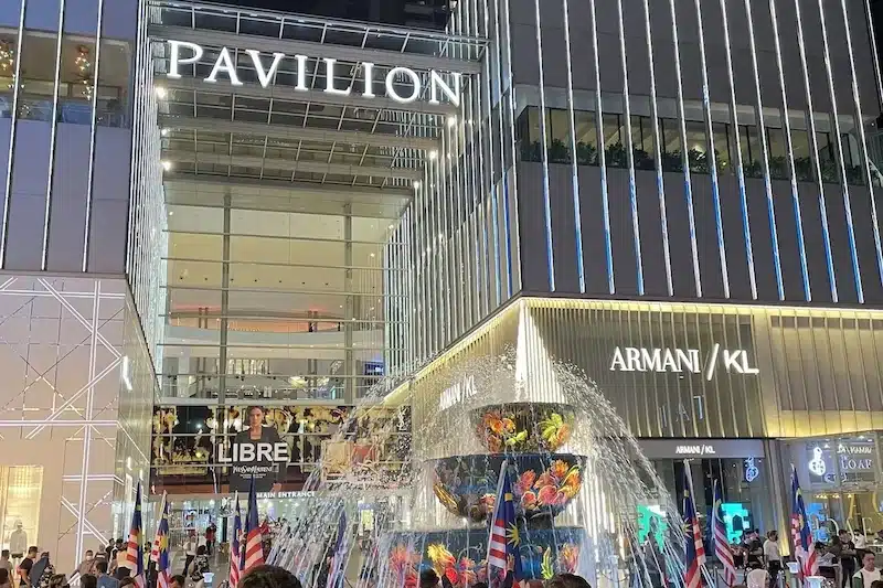 The illuminated main entrance of Pavilion Kuala Lumpur shopping mall featuring the famous crystal fountain. Crowds gather near the luxury storefronts like Armani on a busy evening.