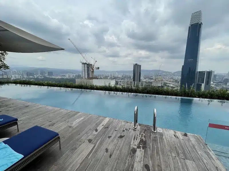 A sleek rooftop infinity pool at the Capri by Fraser hotel with blue lounge chairs. The view overlooks the Kuala Lumpur city skyline, including the Exchange 106 tower, under a cloudy sky.
