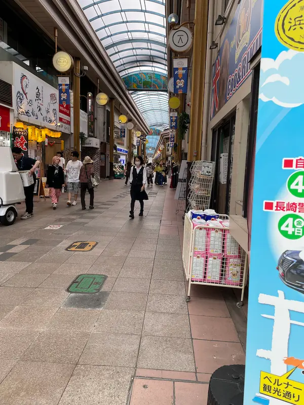Covered shopping street at Hamanmachi Arcade in Nagasaki, Japan, with people walking past restaurants, shops, and colorful signs under a glass roof.