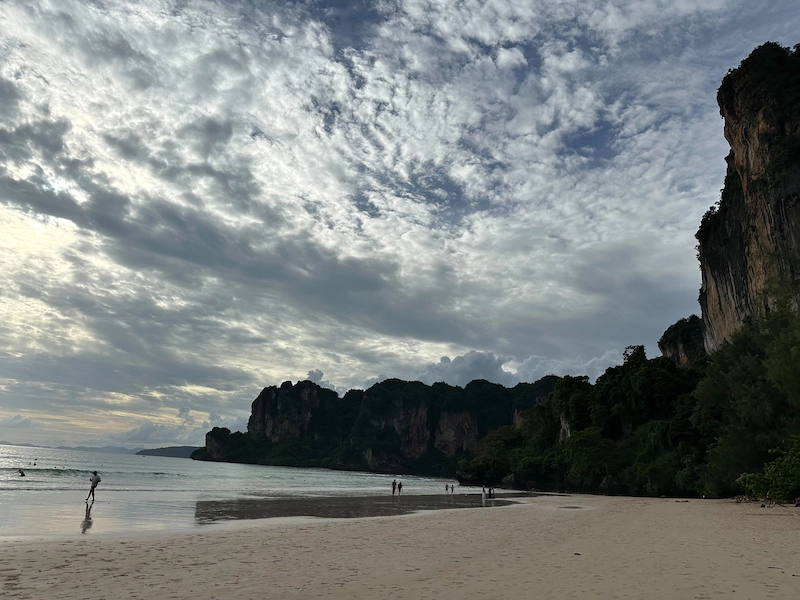 A dramatic view of Railay Beach on a cloudy day, with a few people on the sand dwarfed by the massive, jungle-covered limestone cliffs.