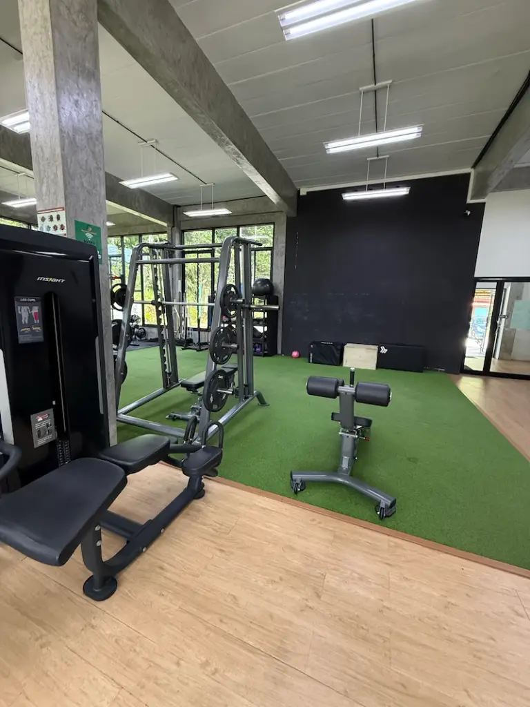 An interior view of Get Well Fitness gym showing a variety of weight training equipment on a light wood floor and a green turf area. In the foreground, there is a preacher curl bench and a hyperextension bench. In the background, there is a power rack with weights, and a black wall at the far end. The gym has an industrial feel with exposed concrete pillars and beams, and large windows letting in natural light.