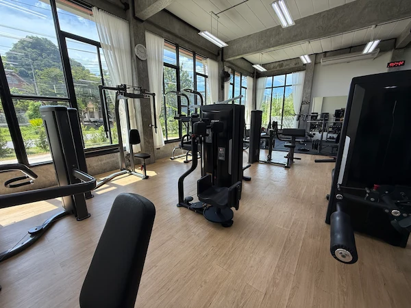 A wide shot of the interior of Get Well Fitness gym, showcasing multiple weight machines and strength training stations. The gym is well-lit with natural light from large windows that offer views of lush greenery outside. The flooring is a light-colored wood laminate, and the ceiling has exposed concrete and fluorescent lighting.