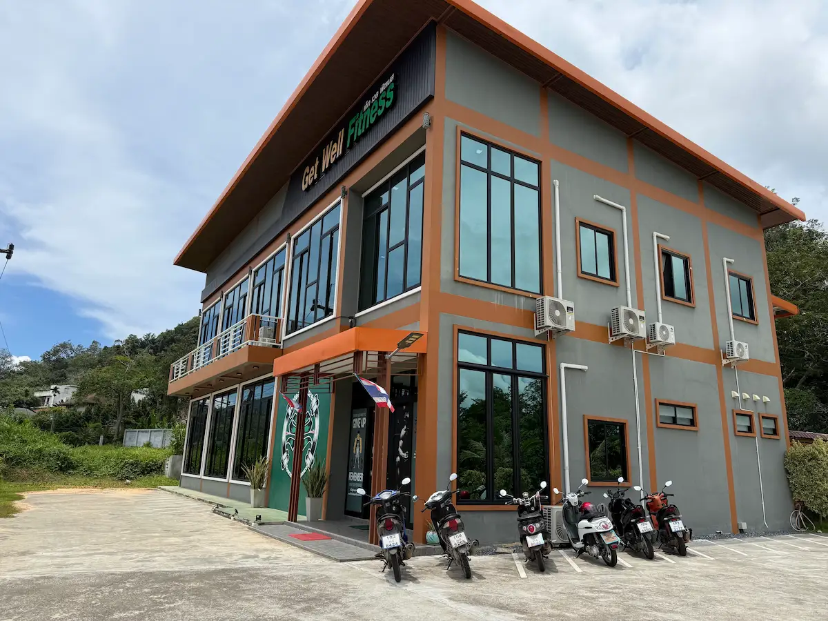 The exterior of the two-story Get Well Fitness gym building in Krabi, Ao Nang, Thailand. It is a modern gray and orange building with large glass windows. The "Get Well Fitness" sign is visible above the entrance. Several scooters are parked in the lot in front of the gym