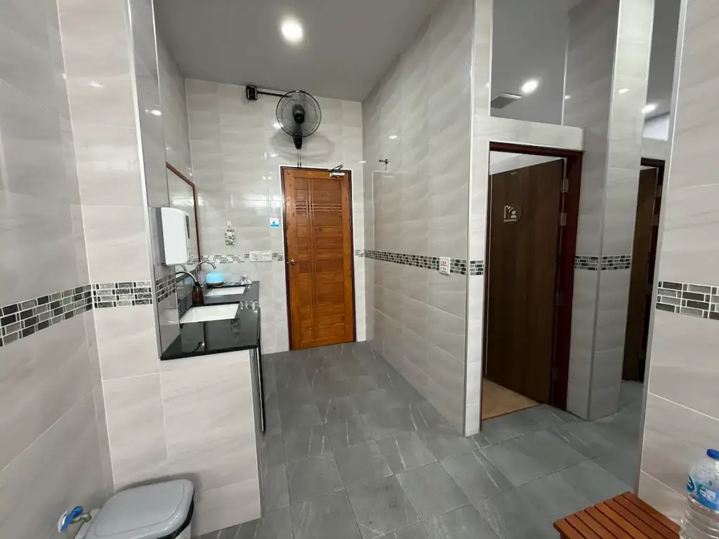 A clean and modern bathroom or changing area at Get Well Fitness gym. The room features light gray tiled walls and floors, a sink with a mirror, and wooden doors leading to what are likely toilets or showers. A wall-mounted fan is visible near the ceiling.