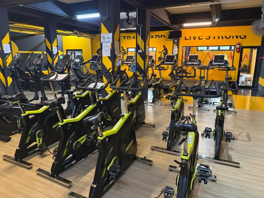 The cardio section of Heroic Fitness Club, featuring rows of modern spin bikes and treadmills. The dominant color scheme is black and yellow, with a bright yellow wall in the background emblazoned with the motivational words "LIVE STRONG" in black.