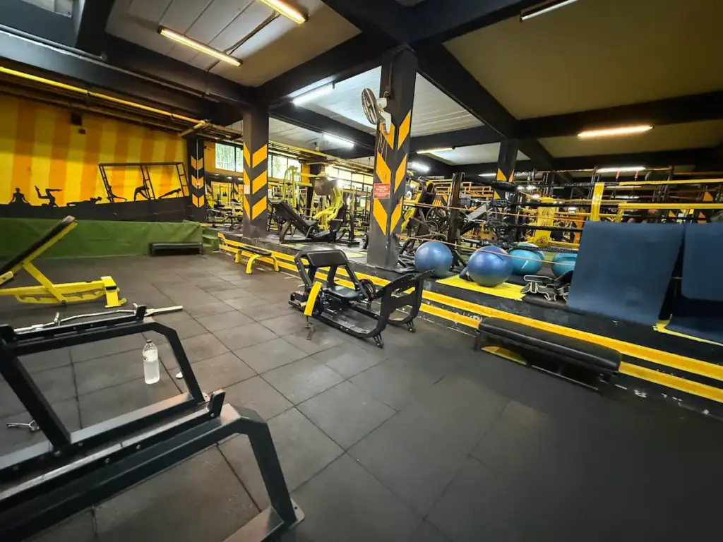 A wide-angle view of the interior of Heroic Fitness Club, showcasing a spacious workout area with black rubber flooring. The gym is filled with a variety of black and yellow weightlifting machines and benches. The walls are yellow with black silhouettes of people exercising, and the structural pillars are black with yellow chevron patterns.
