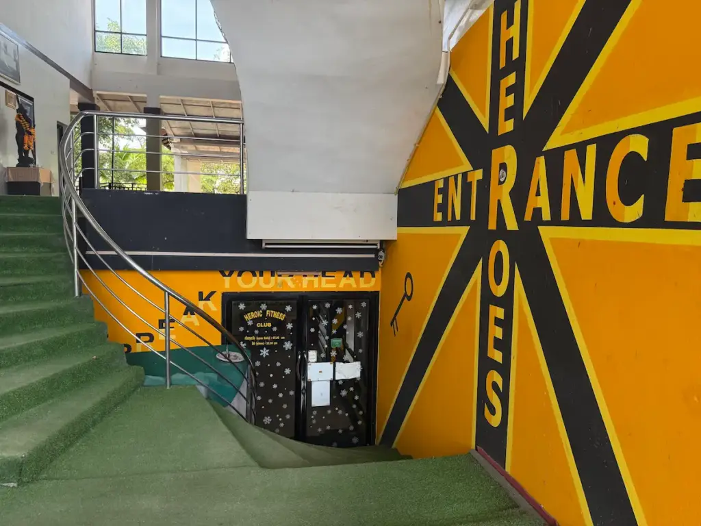 The entrance to the Heroic Fitness Club, with a winding staircase covered in green artificial grass leading down to the club's glass doors. A large, dynamic graphic on the yellow wall reads "HEROES ENTRANCE" in black, designed in a starburst pattern.
