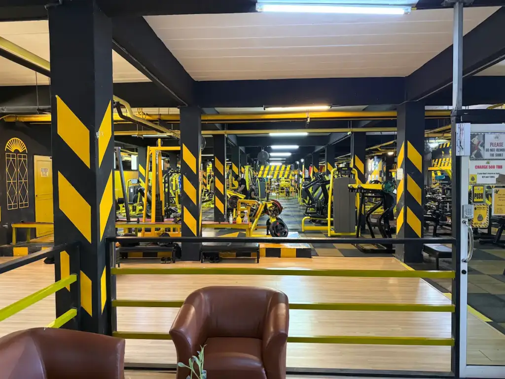A view from a lounge area with brown leather armchairs, looking out over the main floor of Heroic Fitness Club. The gym is expansive, packed with yellow and black exercise equipment under bright lighting. The gym's signature black pillars with yellow stripes are prominent throughout the space.