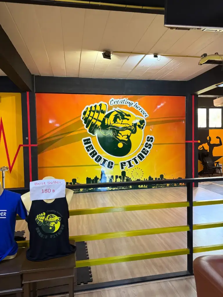A branded wall at Heroic Fitness Club featuring the gym's logo, which depicts a muscular arm with a tiger's head lifting a dumbbell, with the text "Creating heroes" and "HEROIC FITNESS". In front of the wall, inside a training ring with yellow ropes, a black tank top with the gym's logo is displayed on a mannequin with a "Best seller" sign.