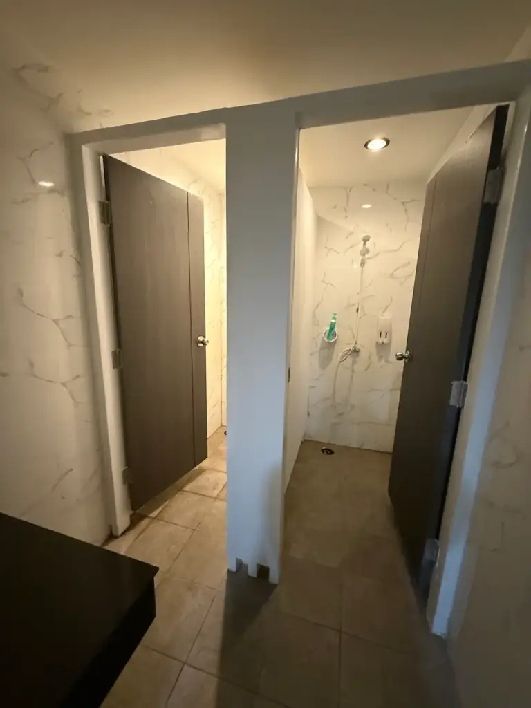 A view of two private shower stalls inside the Heroic Fitness Club. The walls are tiled with a white and gray marble pattern, and the doors are a dark, solid brown. One stall door is partially open, revealing a shower head and soap dispenser inside.