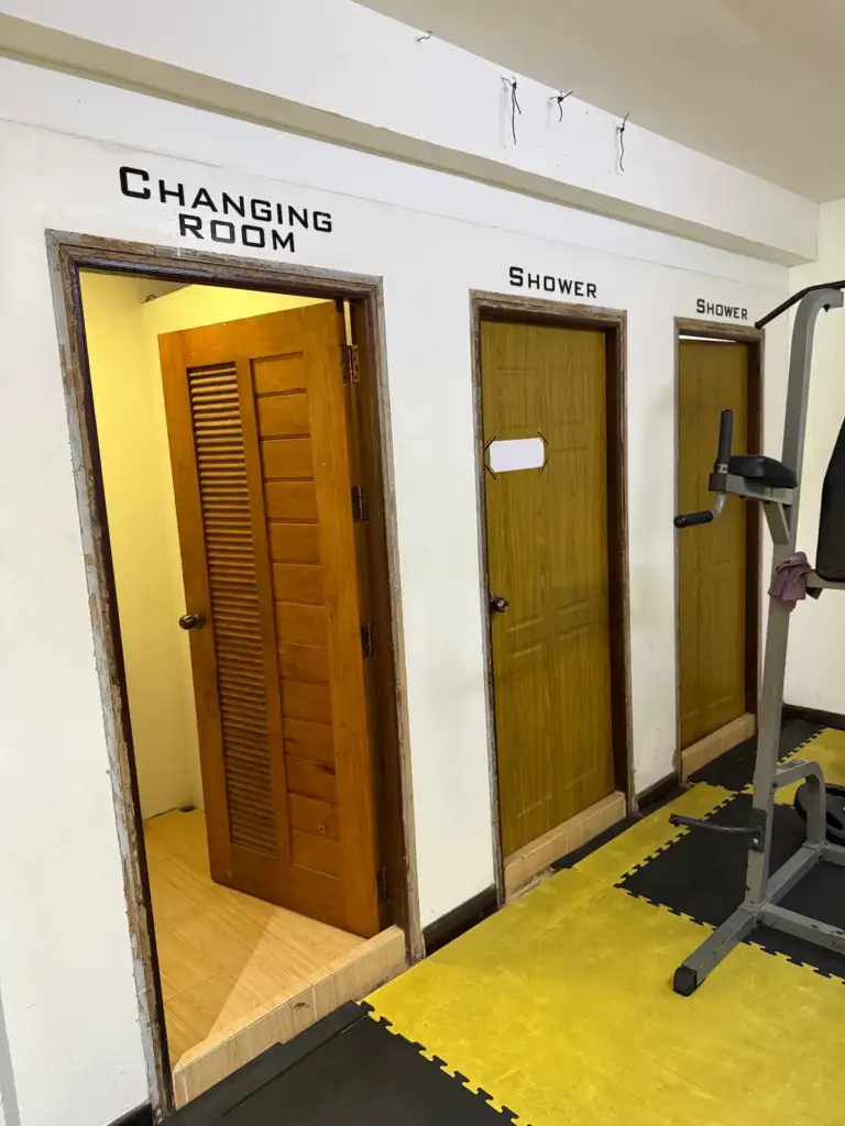 The changing and shower area at PUSH Fitness. A white wall has three wooden doors, with black text above them indicating "CHANGING ROOM" and two "SHOWER"s. The changing room door is open, revealing a lit room inside. The floor is made of black and yellow interlocking safety mats.