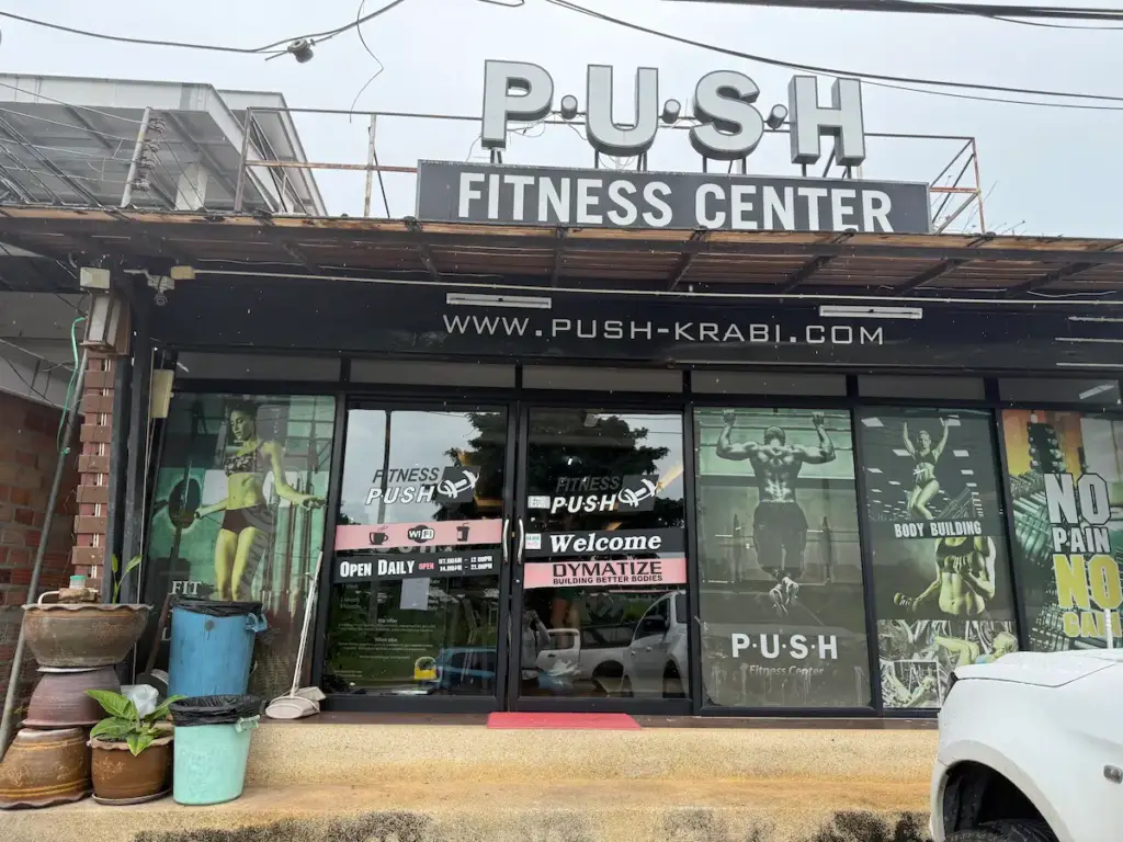 The storefront of PUSH Fitness Center in Ao Nang, Krabi. The entrance features large glass doors and windows adorned with fitness-themed graphics and the gym's logo. A prominent sign above reads "PUSH FITNESS CENTER" in large, bold letters.