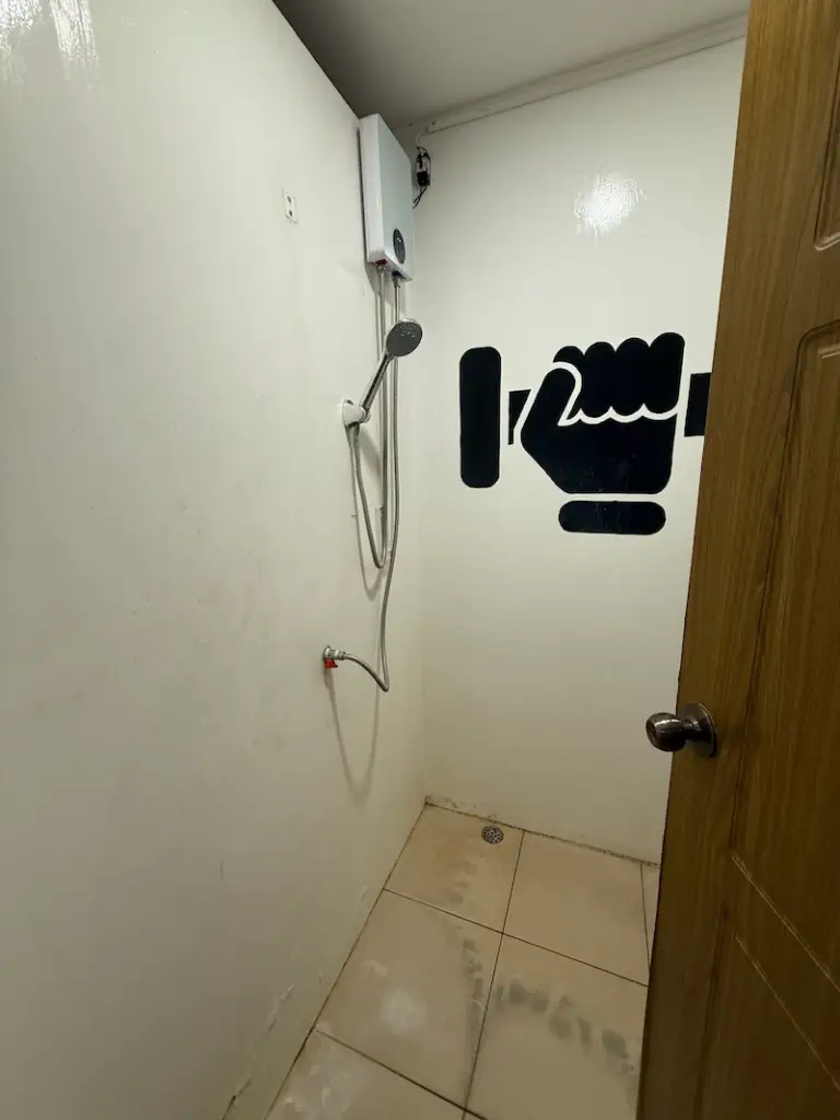 A look inside a basic shower stall at PUSH Fitness. The stall has white walls, a tiled floor, and is equipped with an electric water heater and a handheld shower head. A black vinyl decal of a stylized hand gripping a dumbbell decorates the wall.