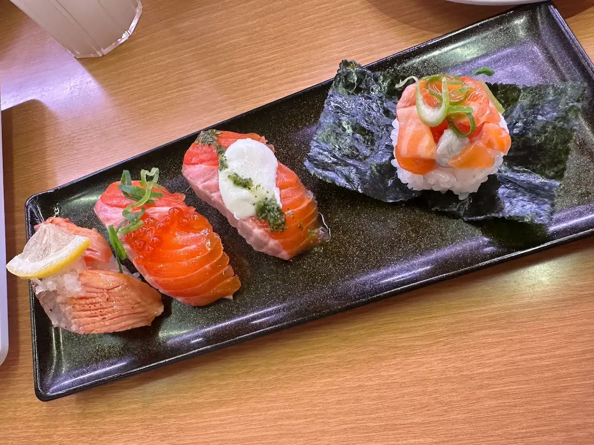 An artistic, long black plate displaying four different types of creatively prepared salmon nigiri, each with unique toppings like fish roe and pesto.