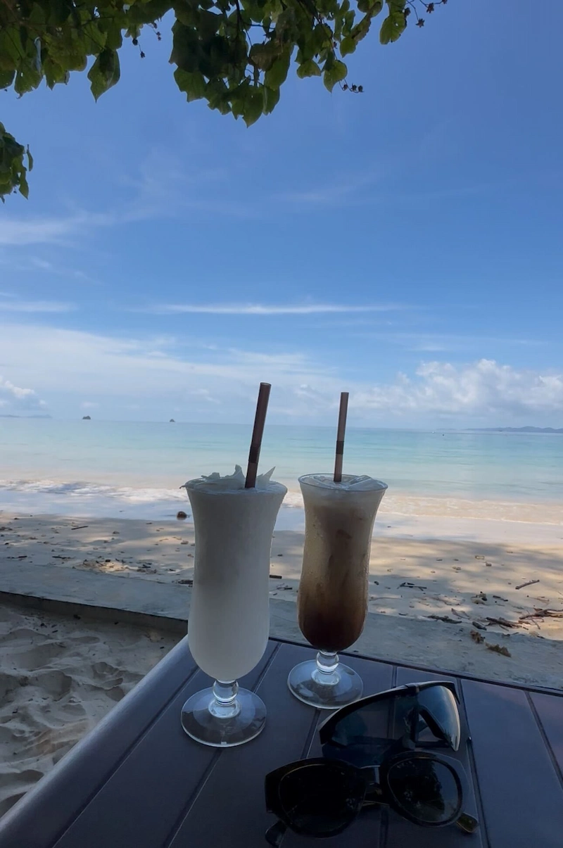 Two tall glasses of iced coffee and a coconut shake sit on a table overlooking a tranquil beach scene, capturing a perfect moment of relaxation in Krabi.