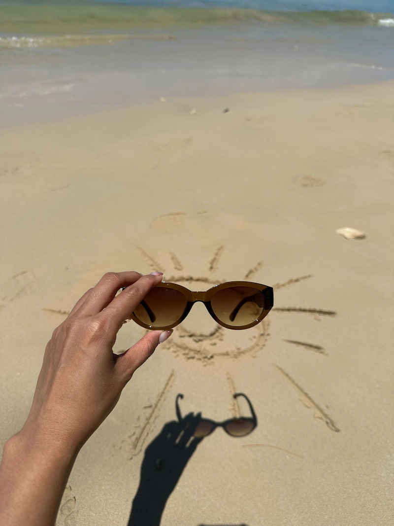 A creative photo of a hand holding a pair of sunglasses over a sun drawn in the sand, with the sunglasses' shadow completing the smiley face.