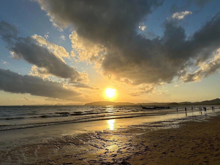 A stunning golden sunset over the ocean at Ao Nang Beach in Krabi. Silhouettes of people and long-tail boats can be seen on the wet sand reflecting the light.