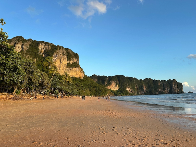 A scenic view of a wide, sandy beach in Krabi during golden hour. People are strolling along the shore, with towering, jungle-covered limestone cliffs lining the coast under a clear blue sky.