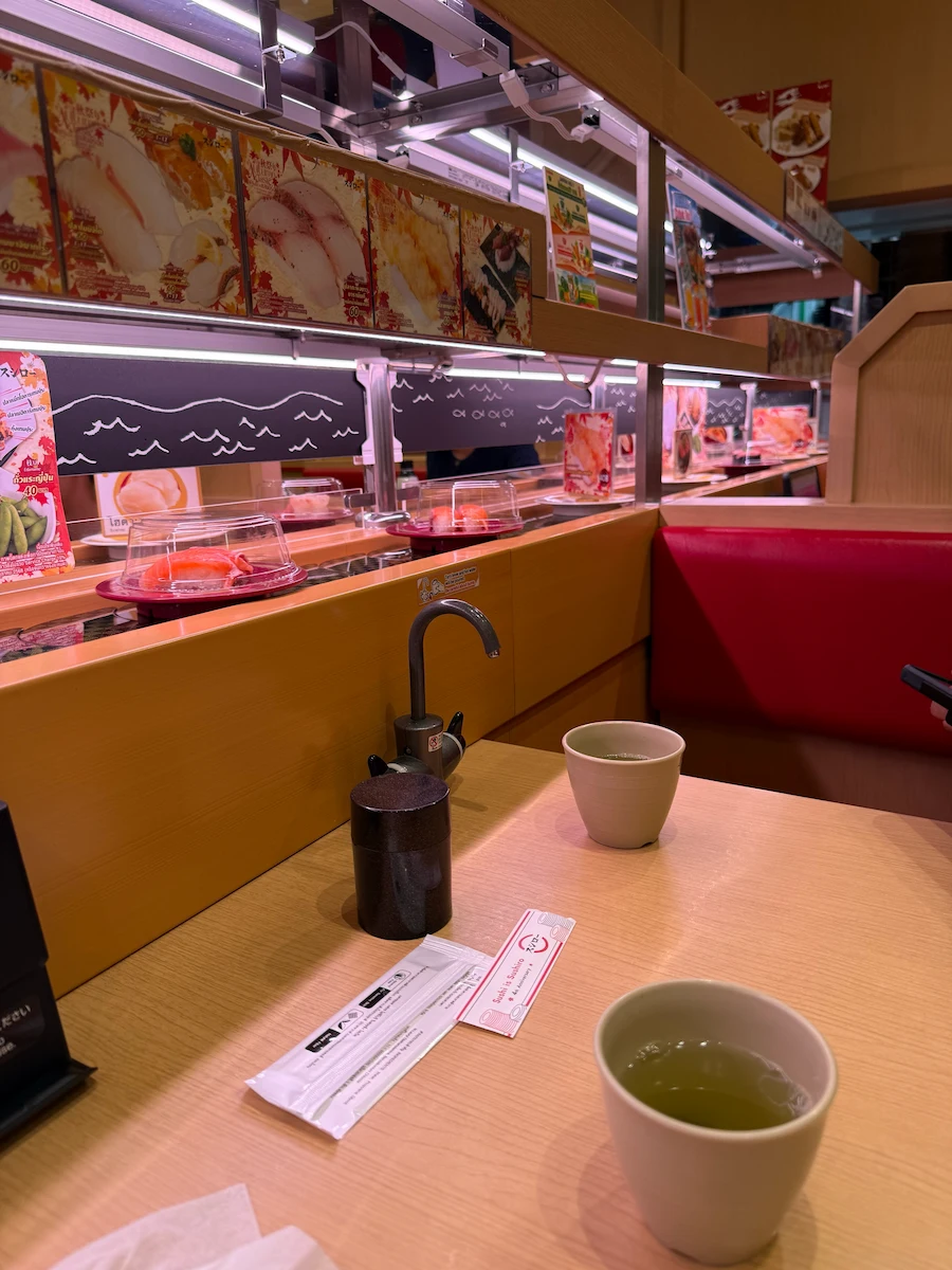 A view from a booth at Sushiro, a conveyor belt sushi restaurant. Two cups of green tea sit on the table, with the two-tiered conveyor belt full of sushi plates in the background.