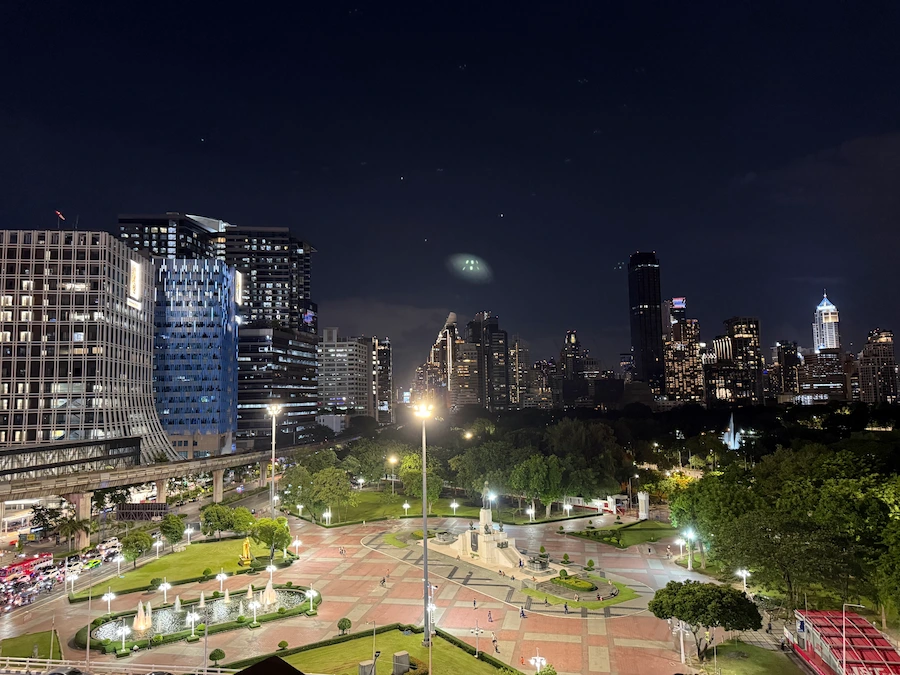 An elevated night view from the mall looking directly over the vast, green, and illuminated Lumpini Park. The King Rama VI monument is visible in the center of the park square.