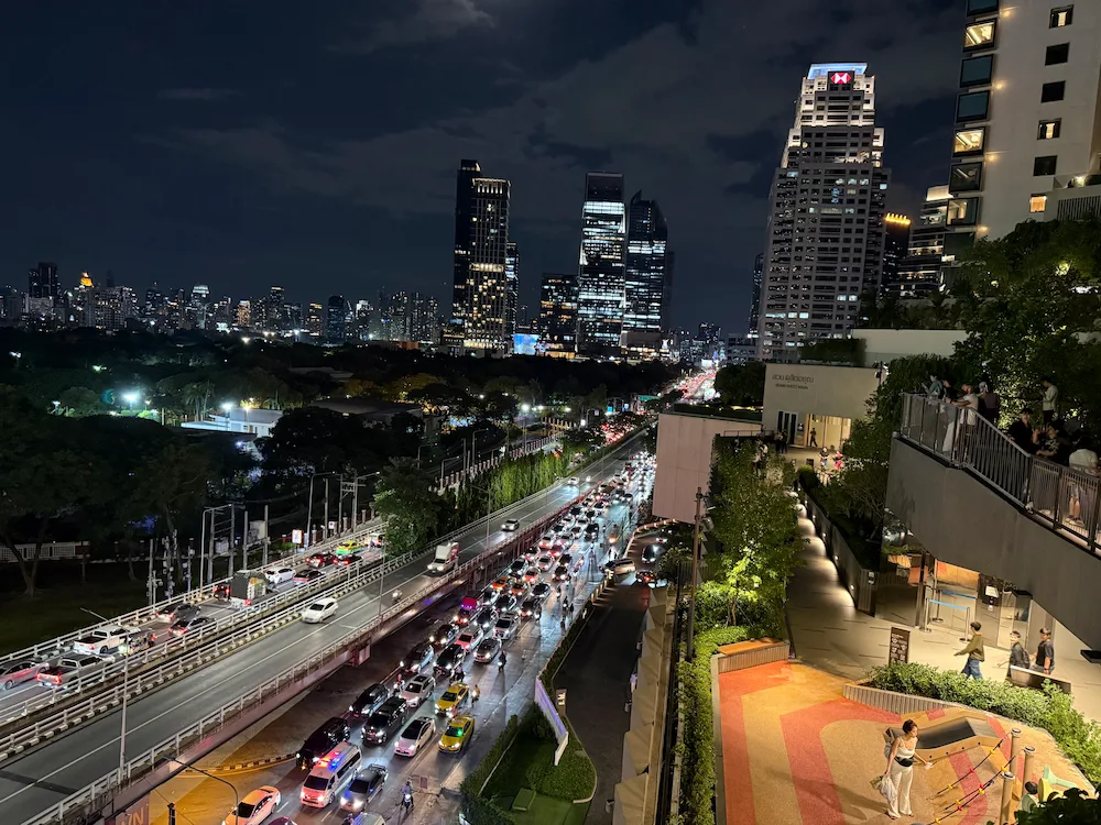 A nighttime view from an outdoor terrace at Dusit Central Park, looking down at the bright streams of traffic on Rama IV road. The illuminated skyscrapers of Bangkok create a dramatic backdrop.